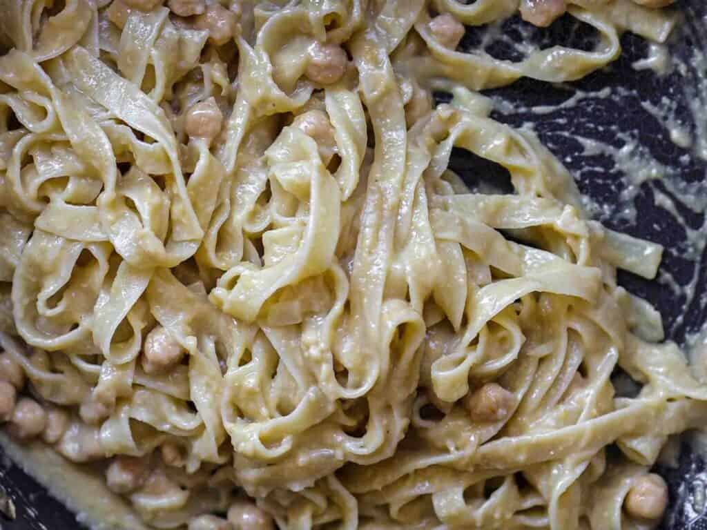 Close-up of creamy pasta mixed with chickpeas, showcasing a rich, sauce-coated texture. The dish has a rustic, homemade appearance with the noodles intertwined and the chickpeas partially visible amidst the sauce.